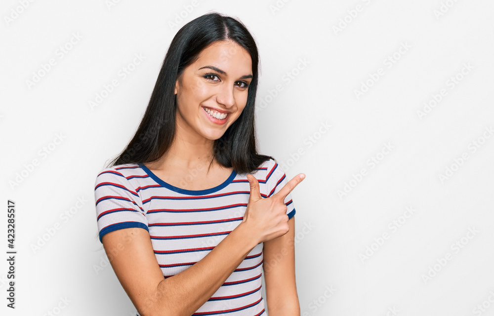 Young hispanic girl wearing casual striped t shirt cheerful with a smile of face pointing with hand and finger up to the side with happy and natural expression on face Young hispanic girl wearing casual striped t shirt cheerful with a smile of face pointing with hand and finger up to the side with happy and natural expression on face