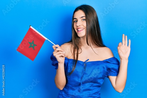 Young brunette teenager holding morocco flag doing ok sign with fingers  smiling friendly gesturing excellent symbol