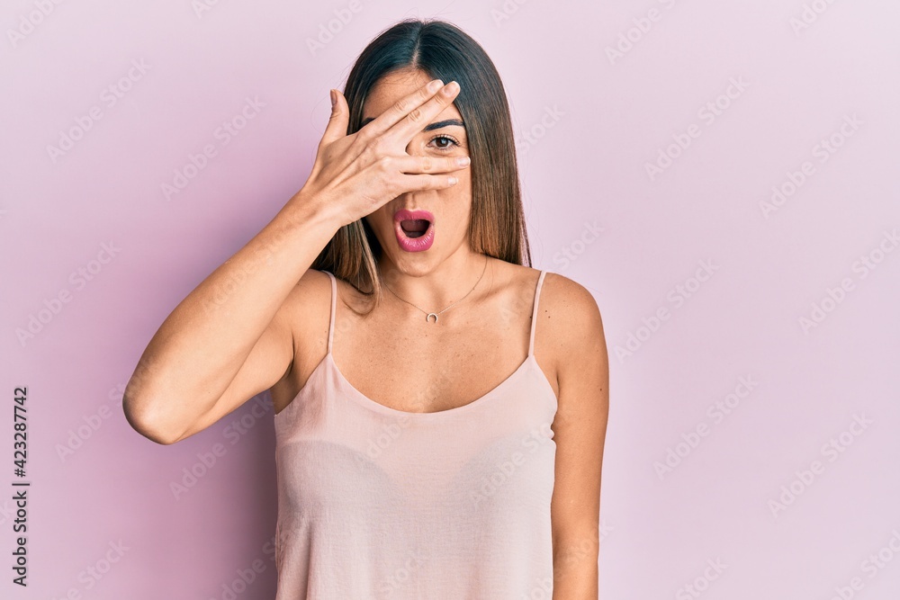 Fototapeta premium Young hispanic woman wearing casual sleeveless t shirt peeking in shock covering face and eyes with hand, looking through fingers afraid