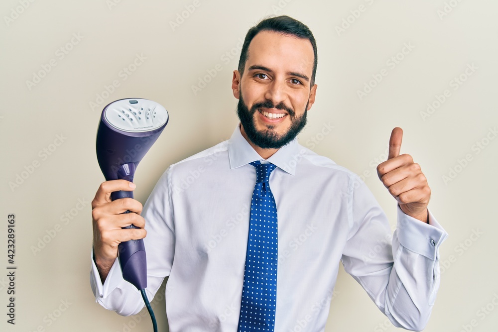Young business man with beard holding electric steam iron smiling happy and positive, thumb up doing excellent and approval sign Young business man with beard holding electric steam iron smiling happy and positive, thumb up doing excellent and approval sign