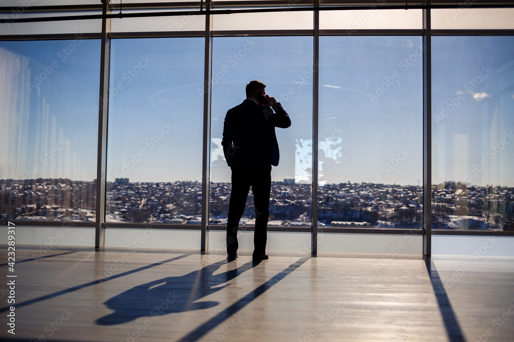 Rear view of a businessman looking out of a large window overlooking the city. He has a phone in his hands. Horizontal view. Rear view of a businessman looking out of a large window overlooking the city. He has a phone in his hands. Horizontal view.
