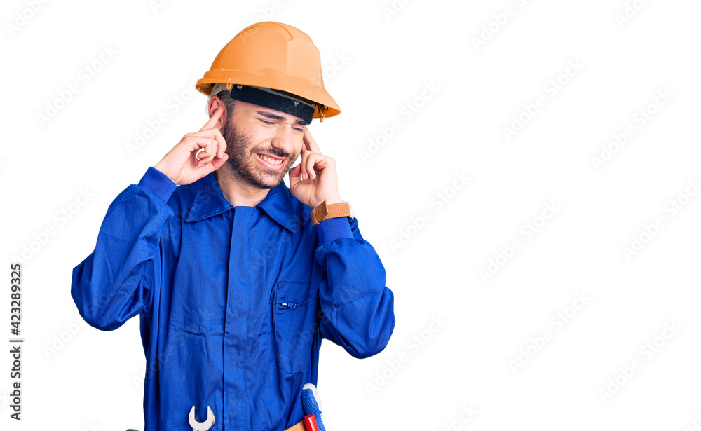 Young hispanic man wearing worker uniform covering ears with fingers with annoyed expression for the noise of loud music. deaf concept.