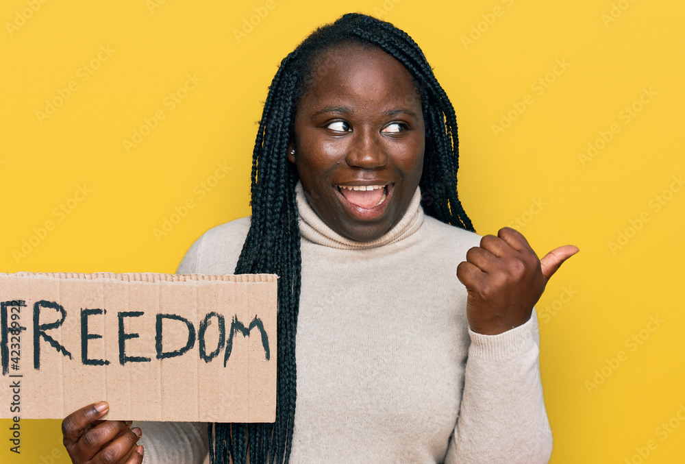 Young black woman with braids holding freedom banner pointing thumb up to the side smiling happy with open mouth Young black woman with braids holding freedom banner pointing thumb up to the side smiling happy with open mouth