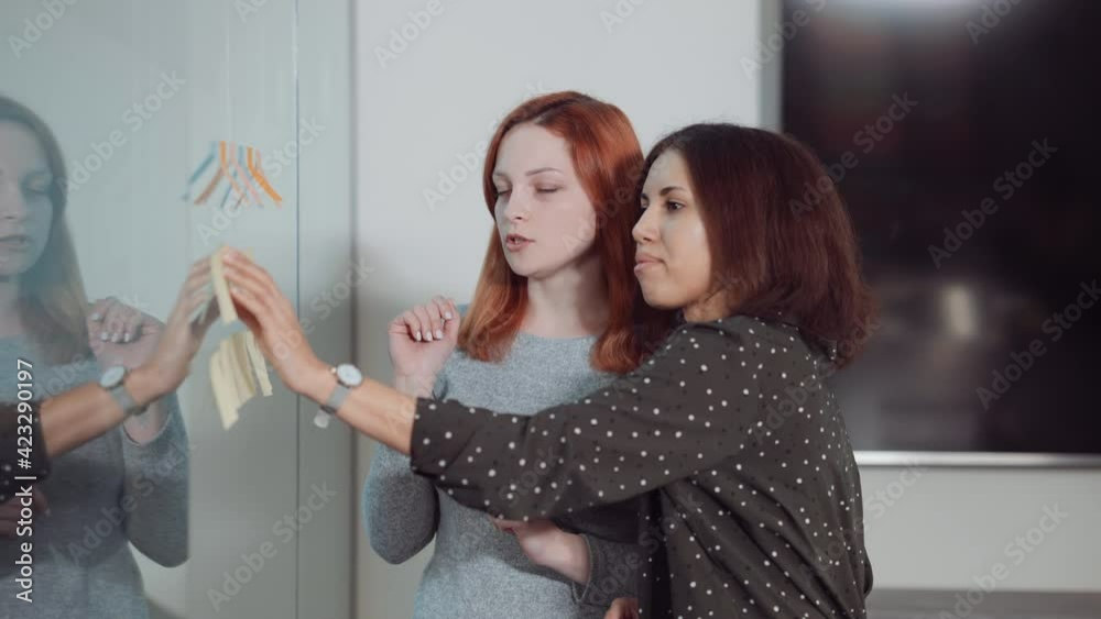 Multiethnic women standing by Scrum board and arranging sticky notes on glass wall. Project managers checking tasks and controlling workflow. Concept of planning