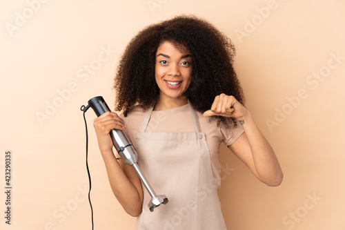 Young african american woman using hand blender isolated on beige background proud and self-satisfied