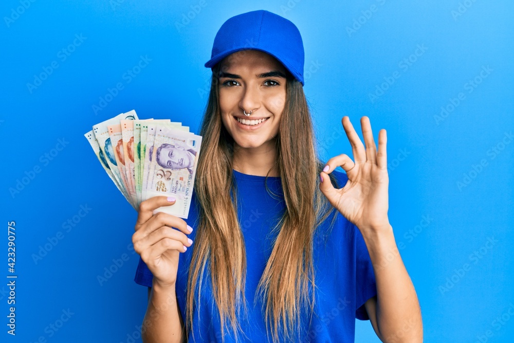 Young hispanic woman wearing delivery uniform and cap holding singapore dollars doing ok sign with fingers, smiling friendly gesturing excellent symbol Young hispanic woman wearing delivery uniform and cap holding singapore dollars doing ok sign with fingers, smiling friendly gesturing excellent symbol