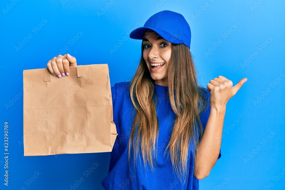Young hispanic woman holding take away paper bag pointing thumb up to the side smiling happy with open mouth Young hispanic woman holding take away paper bag pointing thumb up to the side smiling happy with open mouth