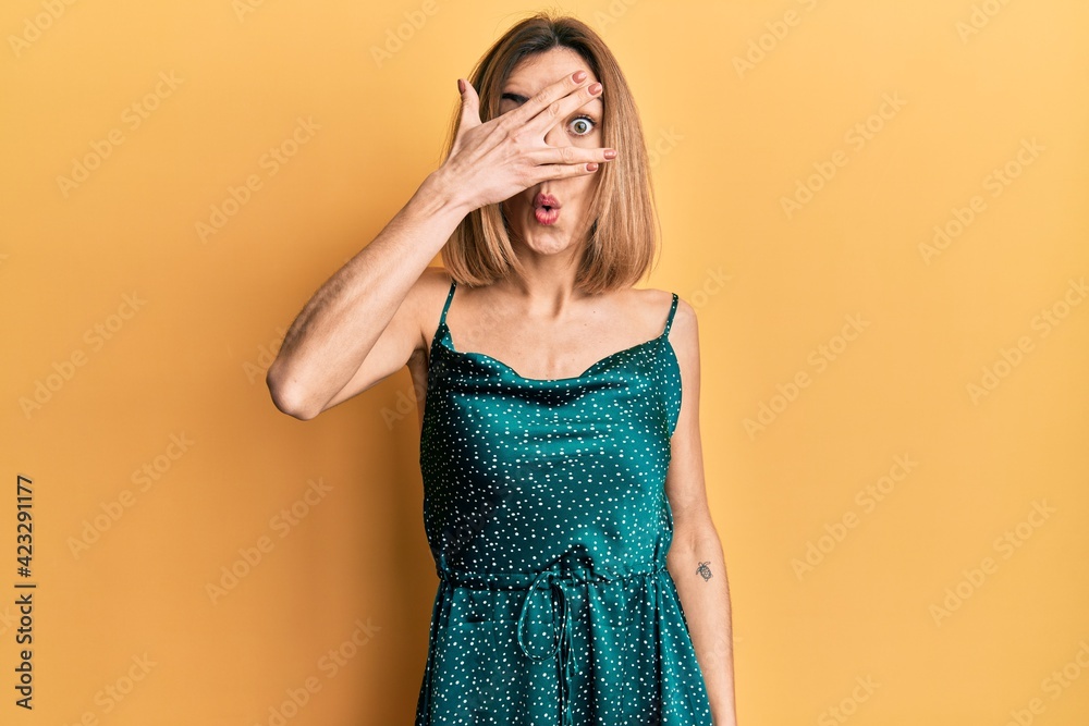 Young caucasian blonde woman wearing elegant party dress peeking in shock covering face and eyes with hand, looking through fingers afraid Young caucasian blonde woman wearing elegant party dress peeking in shock covering face and eyes with hand, looking through fingers afraid