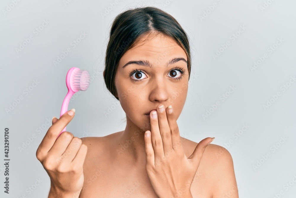 Young brunette woman using facial exfoliating brush covering mouth with hand, shocked and afraid for mistake. surprised expression Young brunette woman using facial exfoliating brush covering mouth with hand, shocked and afraid for mistake. surprised expression