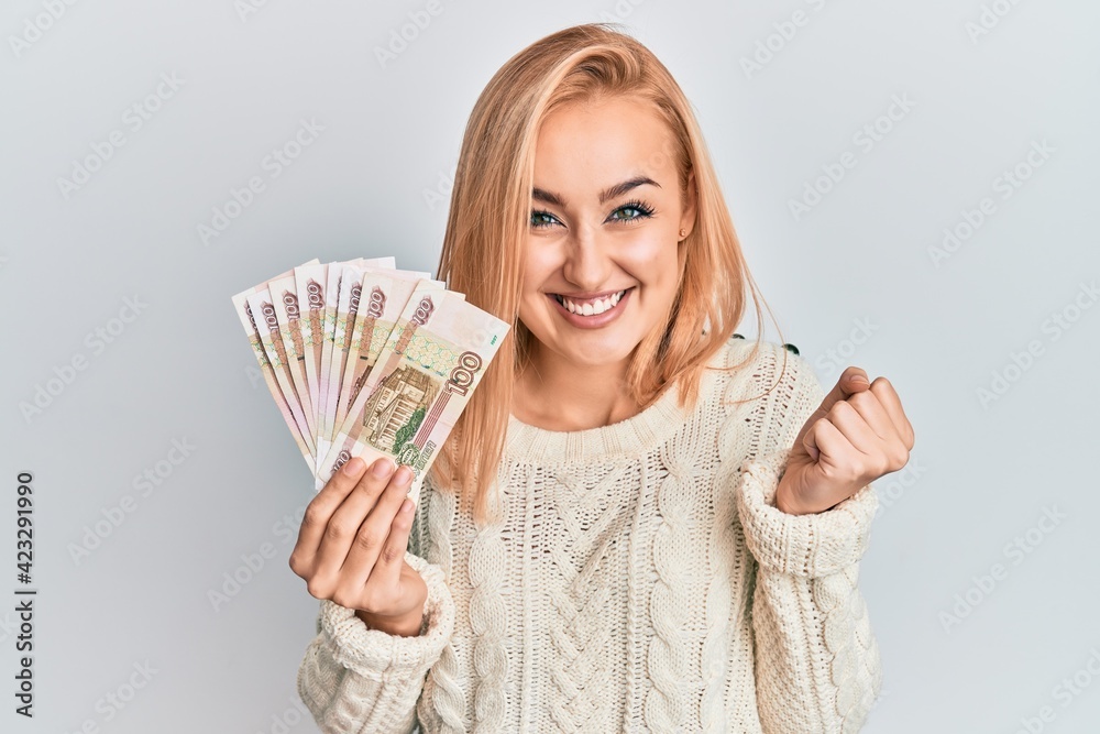 Beautiful caucasian woman holding 100 russian ruble banknotes screaming proud, celebrating victory and success very excited with raised arm Beautiful caucasian woman holding 100 russian ruble banknotes screaming proud, celebrating victory and success very excited with raised arm