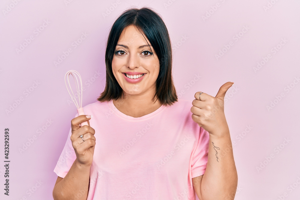 Young hispanic woman cooking using baker whisk smiling happy and positive, thumb up doing excellent and approval sign Young hispanic woman cooking using baker whisk smiling happy and positive, thumb up doing excellent and approval sign