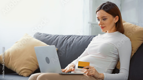 pretty  pregnant woman holding credit card while using laptop on couch