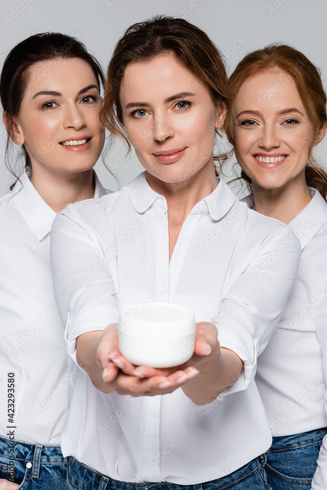 Smiling women looking at camera near friend with jar of cosmetic cream on blurred foreground isolated on grey Smiling women looking at camera near friend with jar of cosmetic cream on blurred foreground isolated on grey