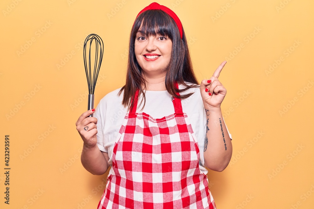 Young plus size woman wearing apron holding whisk smiling happy pointing with hand and finger to the side Young plus size woman wearing apron holding whisk smiling happy pointing with hand and finger to the side