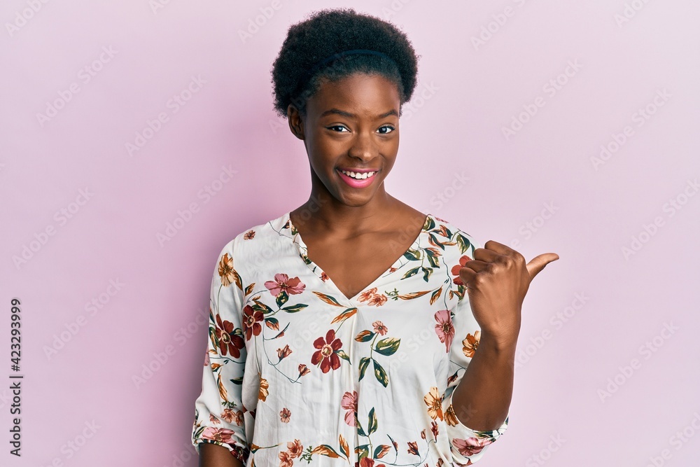 Young african american girl wearing casual clothes smiling with happy face looking and pointing to the side with thumb up. Young african american girl wearing casual clothes smiling with happy face looking and pointing to the side with thumb up.