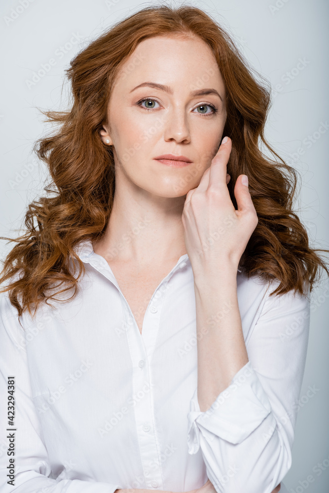Red haired woman with hand near face looking at camera isolated on grey Red haired woman with hand near face looking at camera isolated on grey
