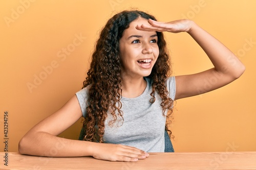 Teenager hispanic girl wearing casual clothes sitting on the table very happy and smiling looking far away with hand over head. searching concept. © Krakenimages.com
