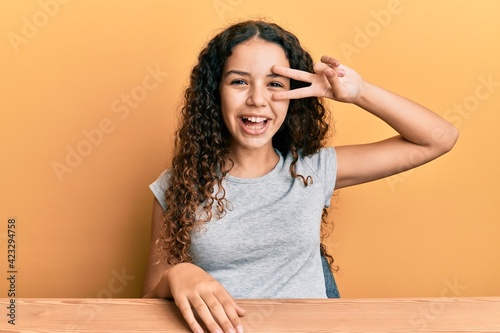 Teenager hispanic girl wearing casual clothes sitting on the table doing peace symbol with fingers over face, smiling cheerful showing victory © Krakenimages.com