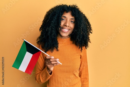 African american woman with afro hair holding kuwait flag looking positive and happy standing and smiling with a confident smile showing teeth
