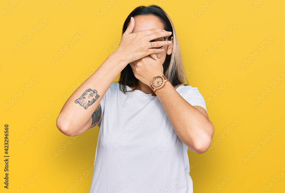 Young hispanic woman wearing casual white t shirt covering eyes and mouth with hands, surprised and shocked. hiding emotion Young hispanic woman wearing casual white t shirt covering eyes and mouth with hands, surprised and shocked. hiding emotion