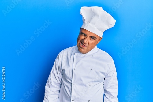 Mature middle east man wearing professional cook uniform and hat winking looking at the camera with sexy expression, cheerful and happy face.