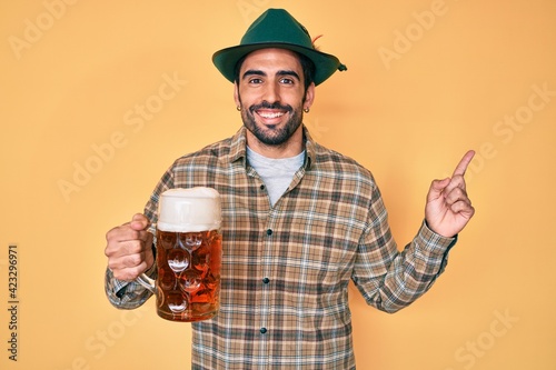 Handsome hispanic man with beard wearing octoberfest hat drinking beer smiling happy pointing with hand and finger to the side