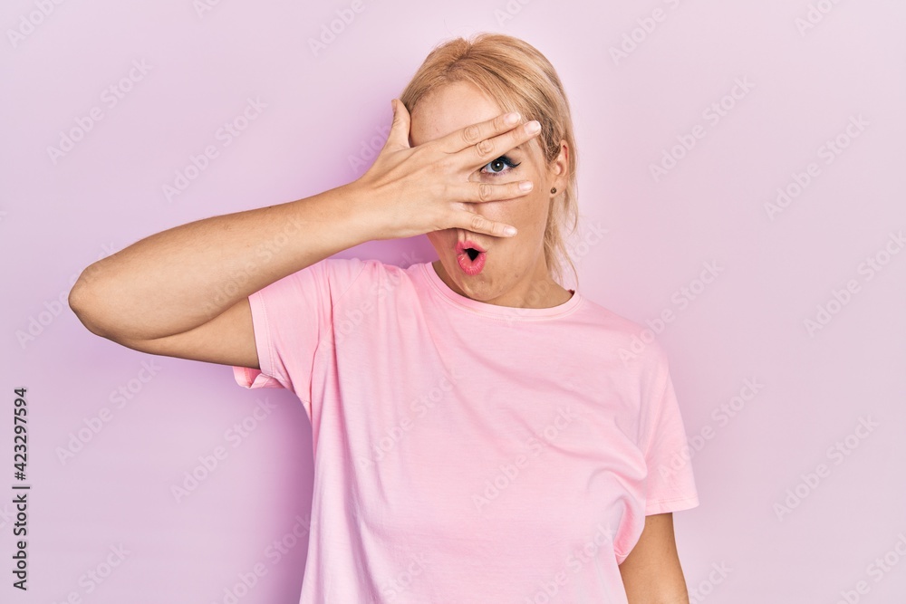 Young blonde woman wearing casual pink t shirt peeking in shock covering face and eyes with hand, looking through fingers with embarrassed expression. Young blonde woman wearing casual pink t shirt peeking in shock covering face and eyes with hand, looking through fingers with embarrassed expression.
