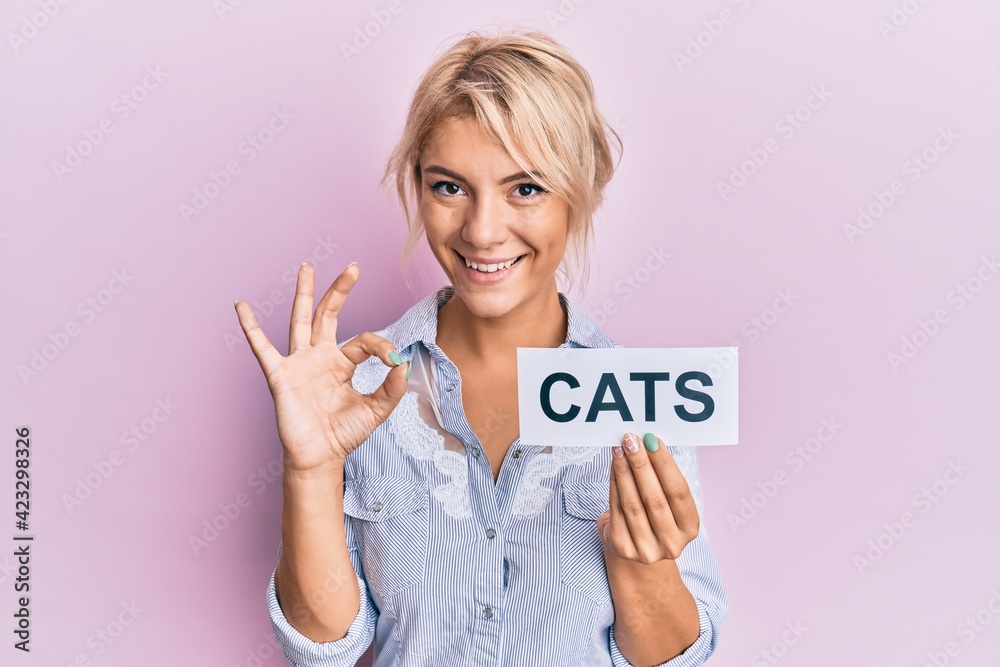 Young blonde girl holding paper with cats word doing ok sign with fingers, smiling friendly gesturing excellent symbol