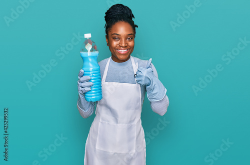 Young african american woman wearing apron holding detergent bottle smiling happy and positive, thumb up doing excellent and approval sign