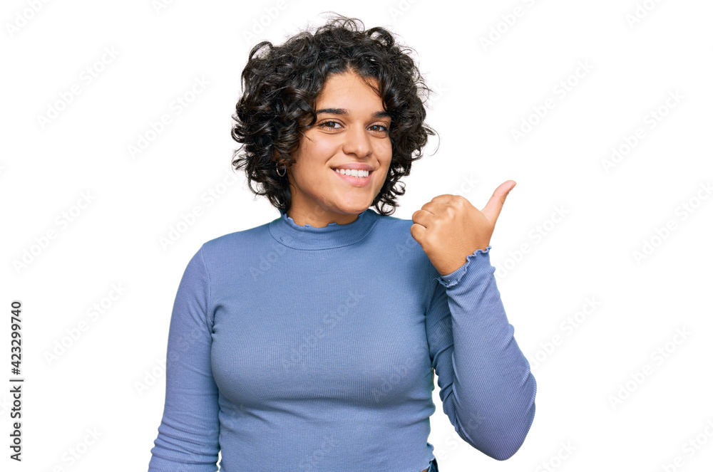 Young hispanic woman with curly hair wearing casual clothes smiling with happy face looking and pointing to the side with thumb up.