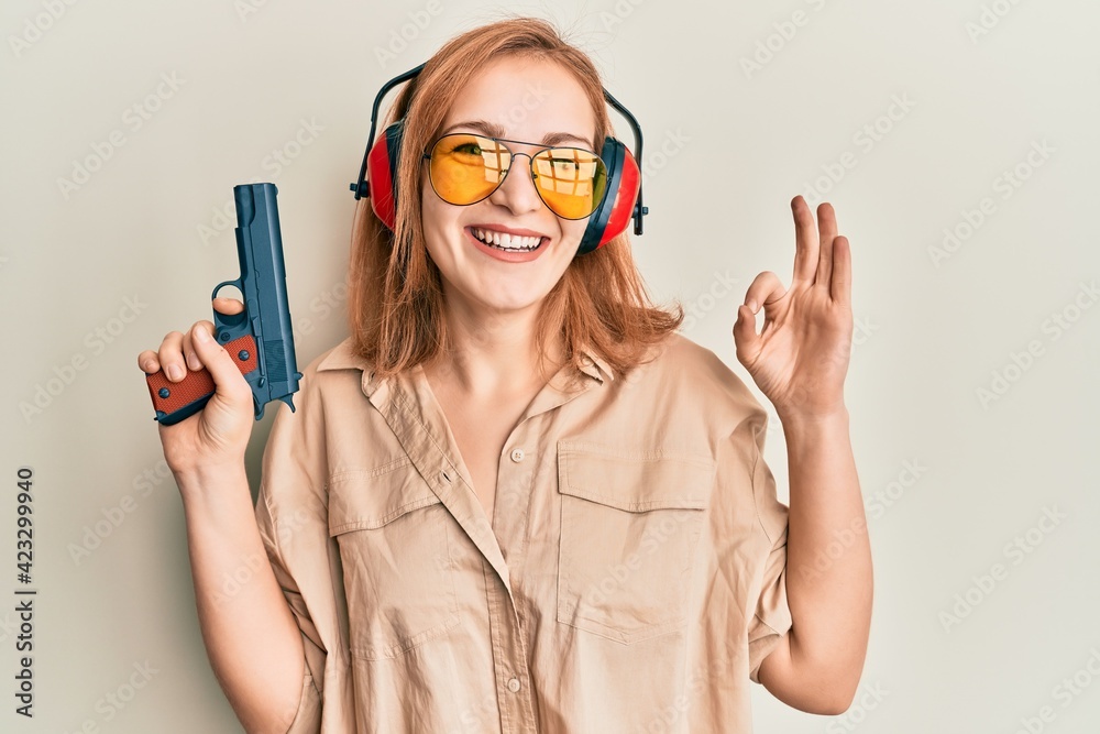 Young caucasian shooter woman using gun doing ok sign with fingers, smiling friendly gesturing excellent symbol