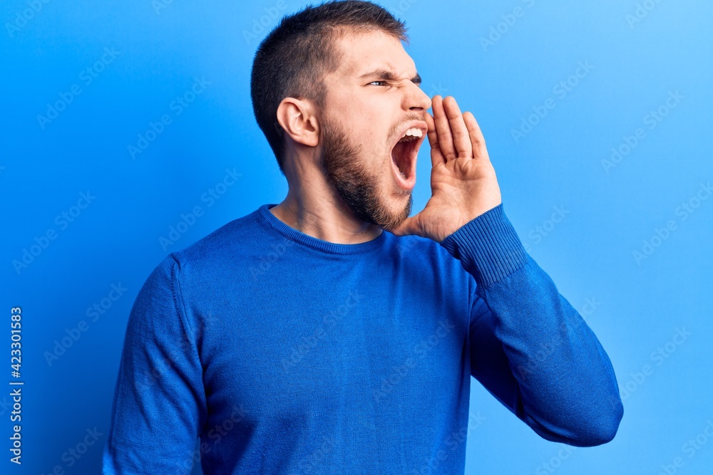 Young handsome man wearing casual sweater shouting and screaming loud to side with hand on mouth. communication concept. Young handsome man wearing casual sweater shouting and screaming loud to side with hand on mouth. communication concept.