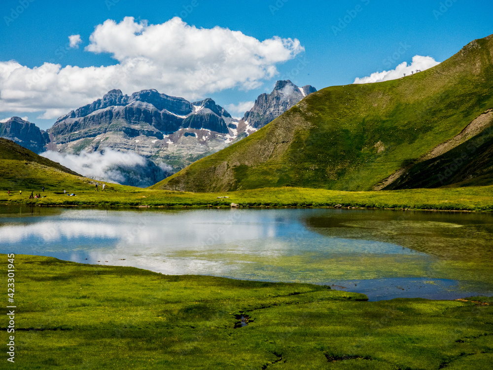 Una laguna de alta montaña y de origen glaciar en un valle colgado rodeado de praderas y con las grandes cumbres nevadas al fondo en los Pirineos españoles
