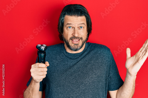Middle age caucasian man holding electric razor machine celebrating victory with happy smile and winner expression with raised hands
