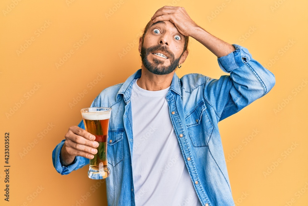 Attractive man with long hair and beard drinking a pint of beer stressed and frustrated with hand on head, surprised and angry face Attractive man with long hair and beard drinking a pint of beer stressed and frustrated with hand on head, surprised and angry face