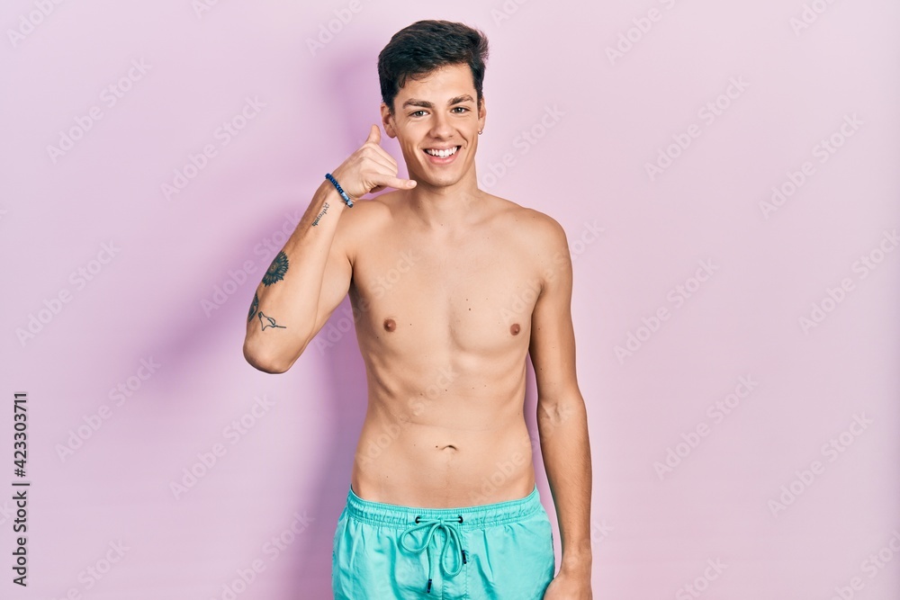 Fototapeta premium Young hispanic man wearing swimwear shirtless smiling doing phone gesture with hand and fingers like talking on the telephone. communicating concepts.