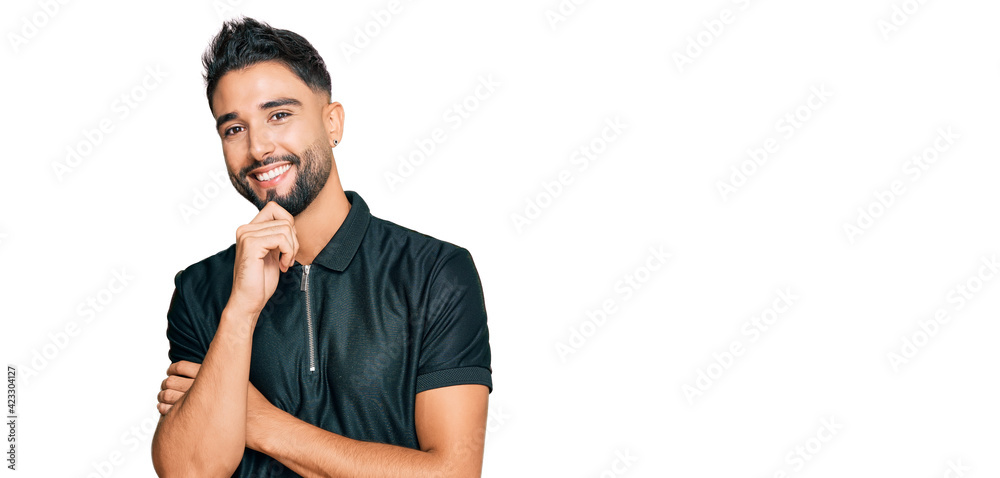 Young man with beard wearing sportswear looking confident at the camera with smile with crossed arms and hand raised on chin. thinking positive. Young man with beard wearing sportswear looking confident at the camera with smile with crossed arms and hand raised on chin. thinking positive.