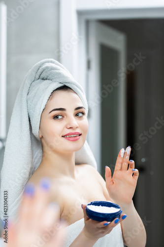 Beauty, skin care and people concept. Smiling young woman applying cream to face and looking to mirror at home bathroom