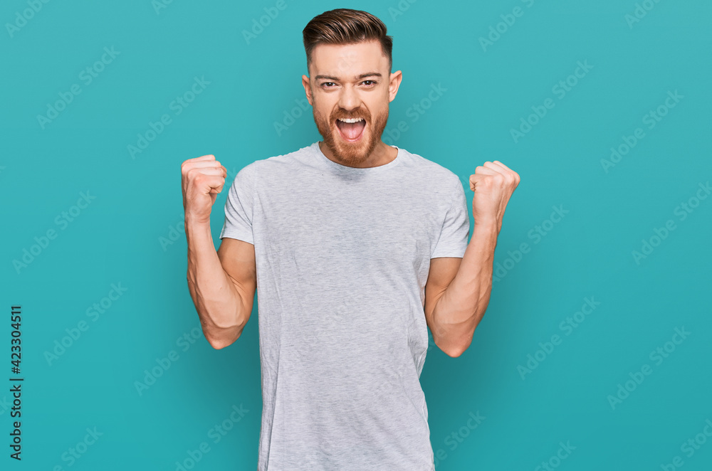 Young redhead man wearing casual grey t shirt celebrating surprised and amazed for success with arms raised and open eyes. winner concept. Young redhead man wearing casual grey t shirt celebrating surprised and amazed for success with arms raised and open eyes. winner concept.