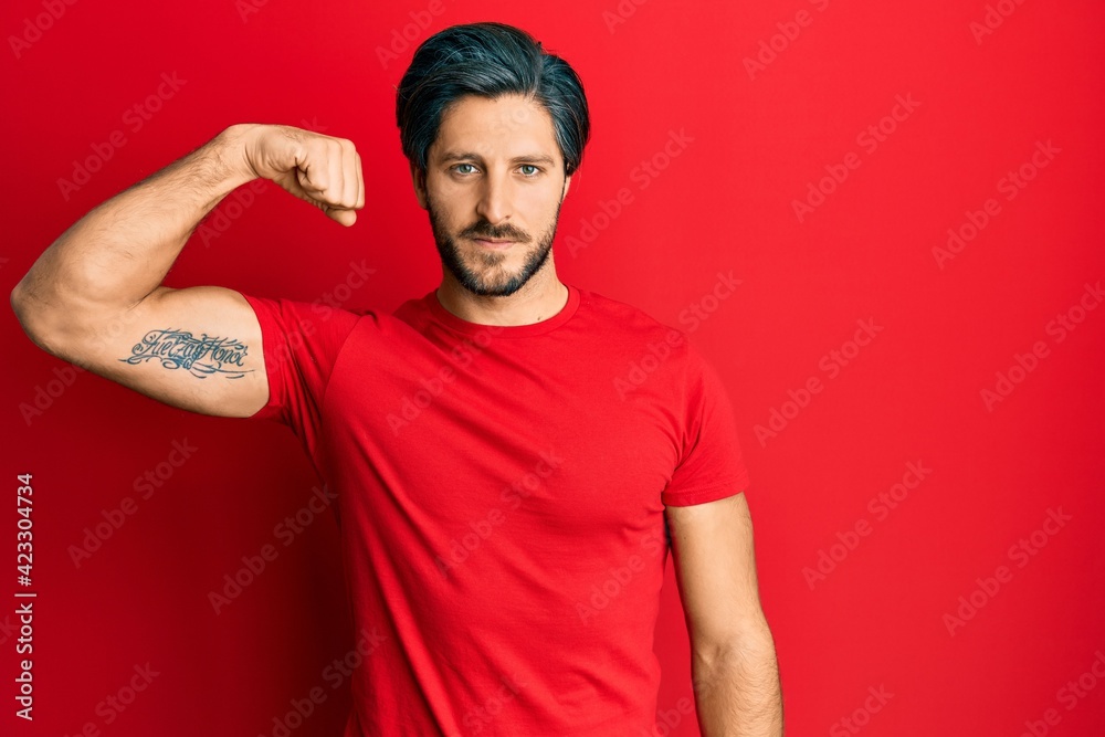 Fototapeta premium Young hispanic man wearing casual red t shirt strong person showing arm muscle, confident and proud of power