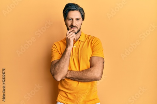 Young hispanic man wearing casual yellow t shirt looking confident at the camera with smile with crossed arms and hand raised on chin. thinking positive. © Krakenimages.com