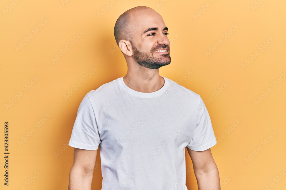Young bald man wearing casual white t shirt looking away to side with smile on face, natural expression. laughing confident. Young bald man wearing casual white t shirt looking away to side with smile on face, natural expression. laughing confident.