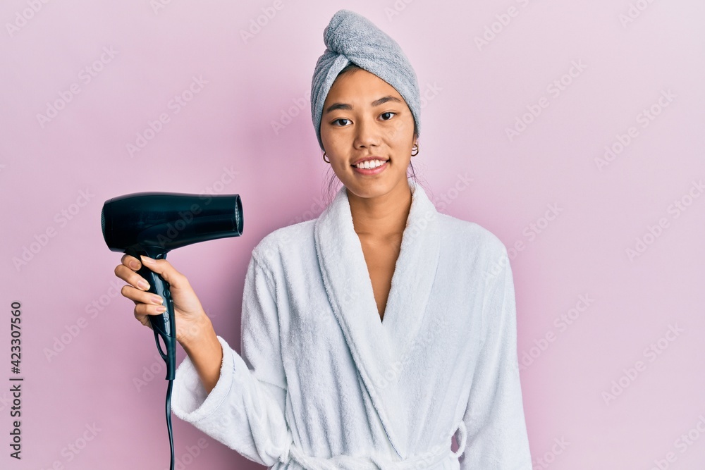 Young chinese woman wearing shower towel cap and bathrobe holding dryer looking positive and happy standing and smiling with a confident smile showing teeth Young chinese woman wearing shower towel cap and bathrobe holding dryer looking positive and happy standing and smiling with a confident smile showing teeth