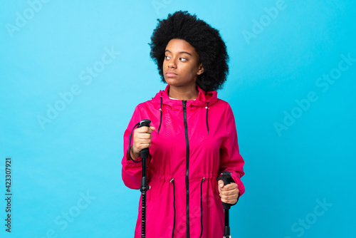 Young Africa American with backpack and trekking poles isolated on blue background having doubts while looking side
