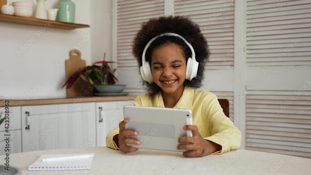 Little African American girl in big white headphones plays a game using a digital tablet. Rest during online learning, break, leisure. A teenage girl sits at a table in a bright kitchen. Slow motion. Little African American girl in big white headphones plays a game using a digital tablet. Rest during online learning, break, leisure. A teenage girl sits at a table in a bright kitchen. Slow motion.