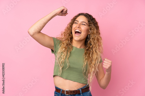 Young blonde woman with curly hair isolated on pink background celebrating a victory