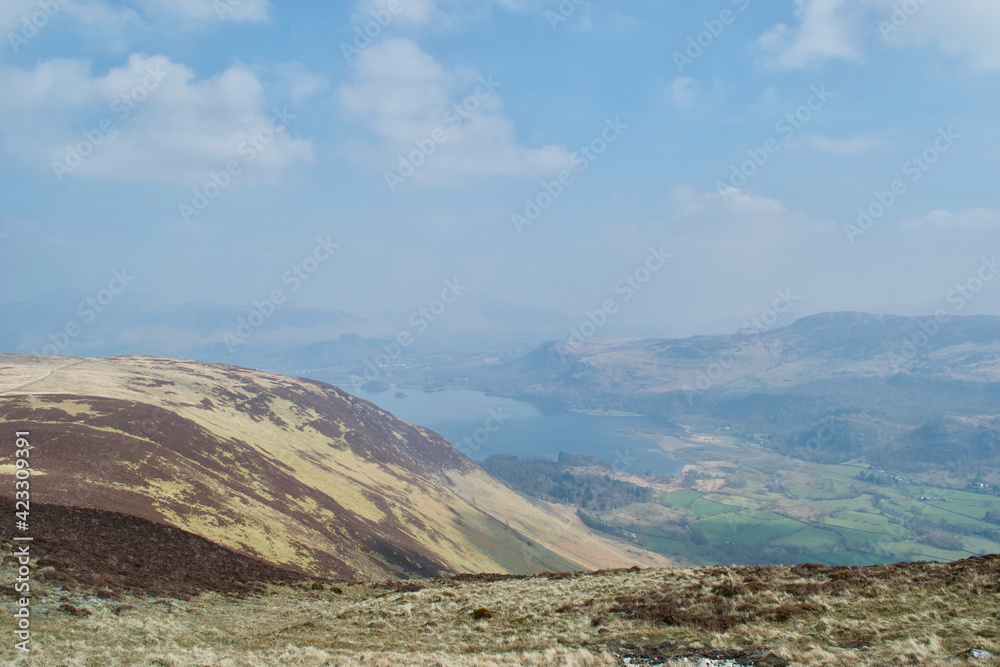 Looking across the yellow side of a mountain top, across a beautiful rural valley to a wide lake in a haze. Misty day viewing Derwent Water, the Lake District, England UK