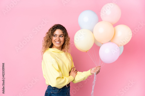 Young blonde woman with curly hair catching many balloons isolated on pink background laughing