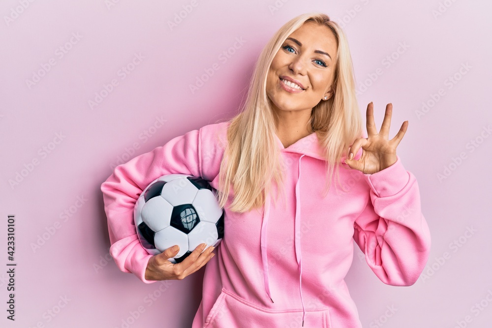 Young blonde woman holding soccer ball doing ok sign with fingers, smiling friendly gesturing excellent symbol Young blonde woman holding soccer ball doing ok sign with fingers, smiling friendly gesturing excellent symbol