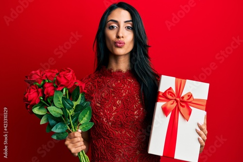 Beautiful hispanic woman holding anniversary gift and roses bouquet looking at the camera blowing a kiss being lovely and sexy. love expression.